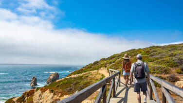 Couple walking on wooden pathway by the ocean. Summer mountain coastal landscape. Garrapata State Park, View from highway 1. Big Sur, California, USA.