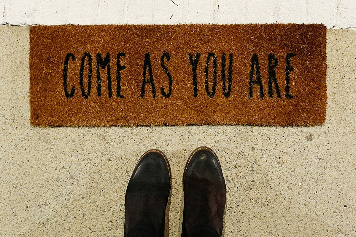 Person standing in front of a doormat that reads "Come As You Are"