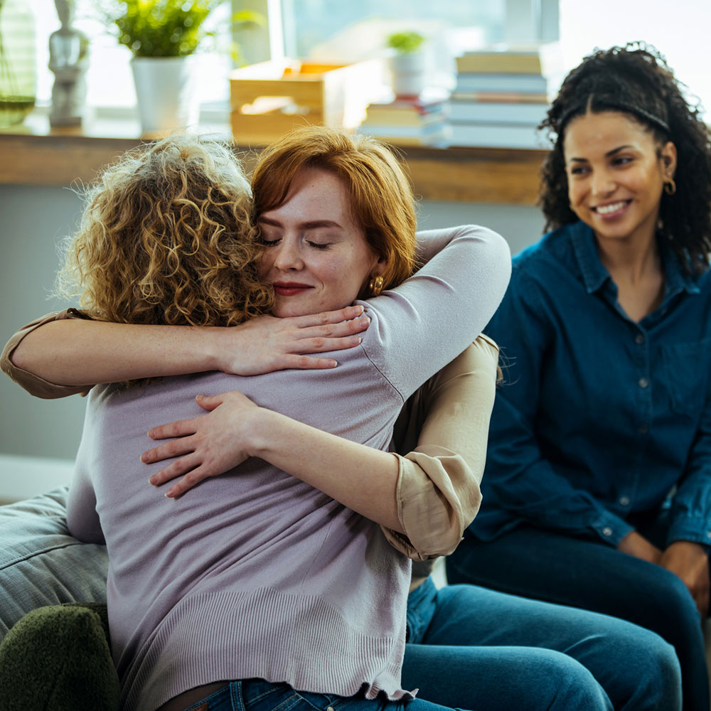 Caring female counselor hugs a female patient during a group therapy session.